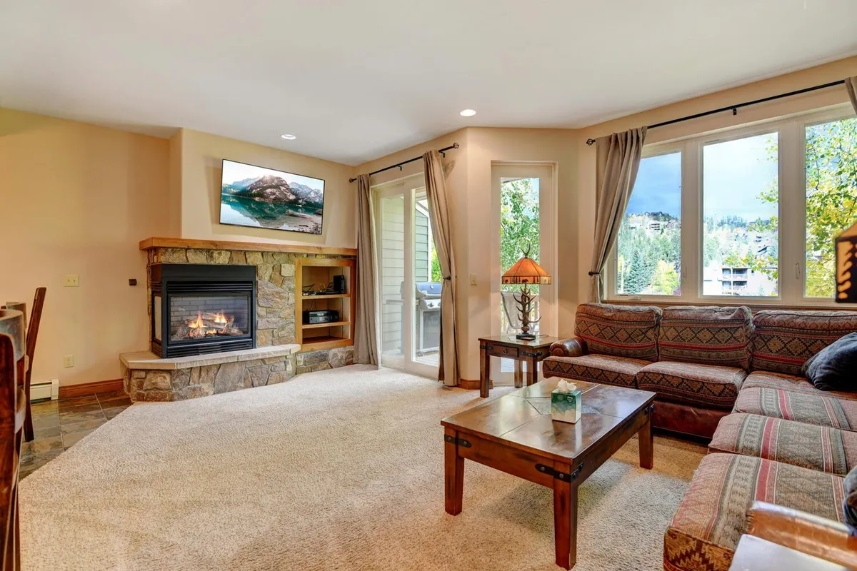 A cozy living room with a plush sofa arranged around a glowing fireplace, large windows that let in abundant natural light, and a modern coffee table. The space feels open and inviting, ideal for relaxing, socializing, or enjoying a movie night on the wall-mounted TV.