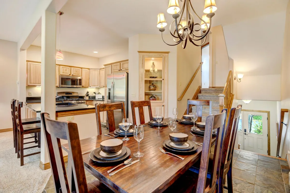 A spacious dining area featuring a wooden dining table under a stylish chandelier, adjacent to a modern kitchen area and an elegant staircase. The layout is open and bright, perfectly suited for shared meals, celebrations, or morning coffee rituals.