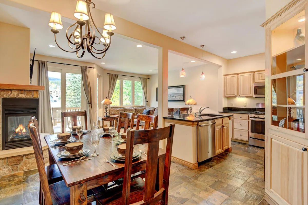 The image showcases a spacious, warmly lit interior featuring a beautifully set dining table, a cozy fireplace, a modern kitchen island, and expansive windows that likely reveal mountain or river views. The inviting scene suggests the perfect setting for memorable family meals, festive gatherings, and relaxed mornings in the heart of Winter Park.