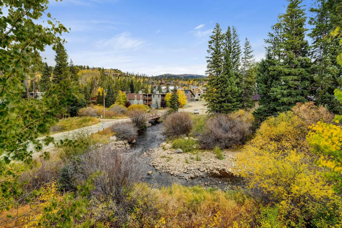 A scenic view of a gentle stream bordered by vibrant autumn foliage and surrounded by trees, with majestic mountains towering in the distance. There are buildings visible further away, suggesting proximity to a charming mountain town.