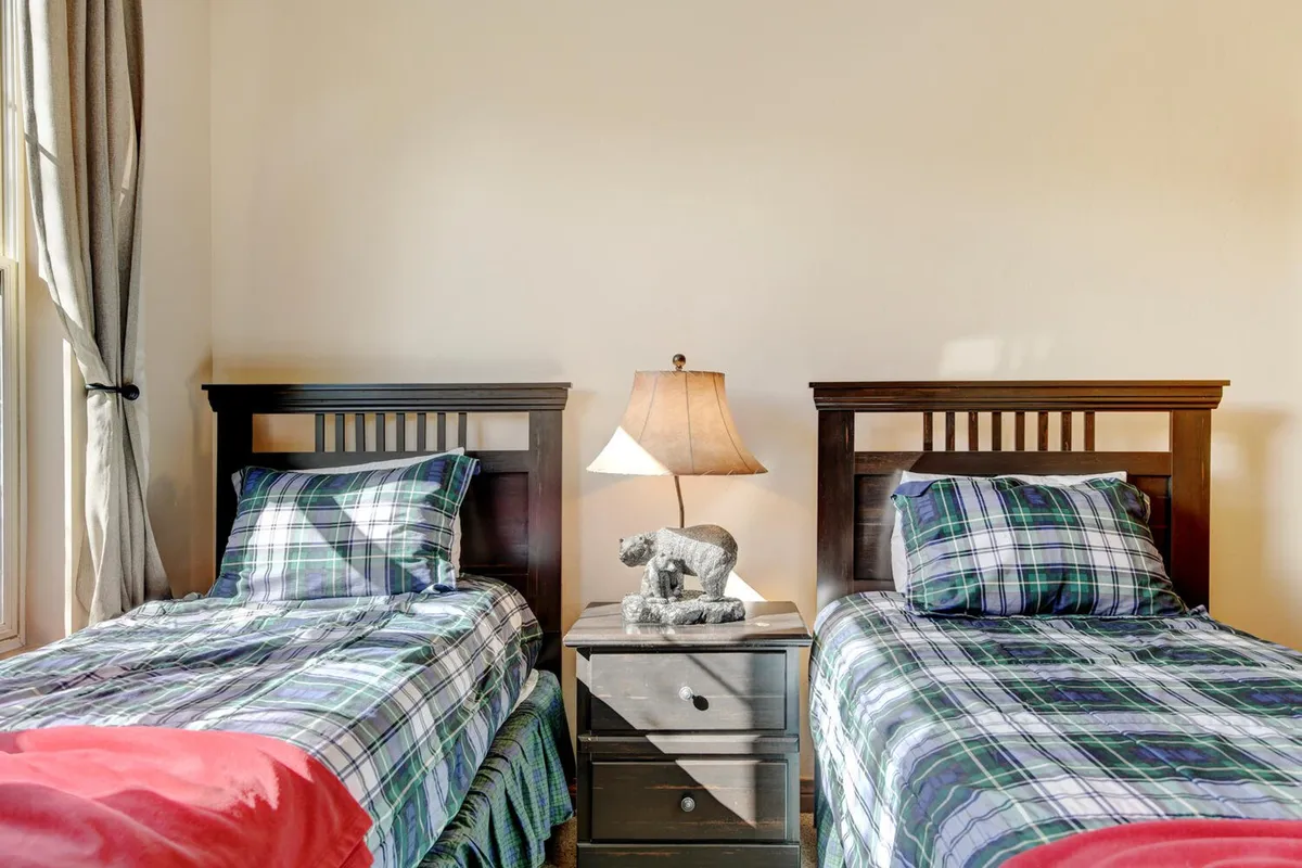 A bright bedroom with two twin beds dressed in plaid bedding, a shared nightstand with a lamp, and a window streaming in natural daylight. The room's layout is versatile, catering to different group needs.