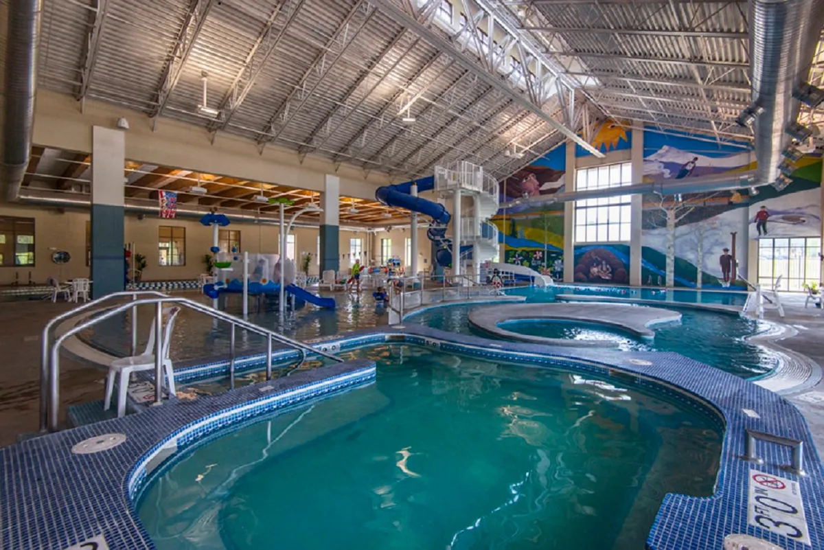 A lively indoor recreation area with water slides, children playing in a pool, colorful murals on the walls, and wavy pool edges. The vibrant atmosphere highlights fun and energetic family activities.